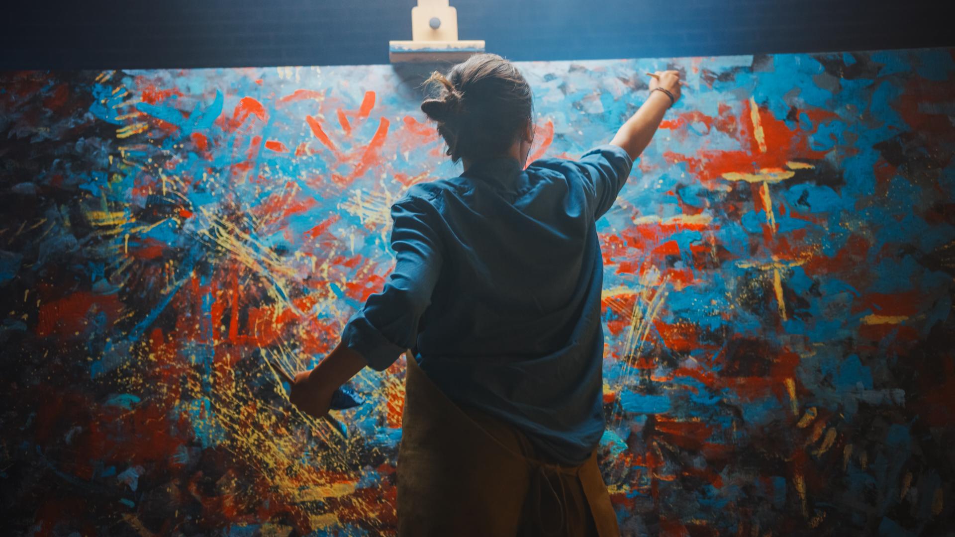 Talented Female Artist Works on Abstract Oil Painting, Using Paint Brush She Creates Modern Masterpiece. Dark and Messy Creative Studio where Large Canvas Stands on Easel Illuminated.